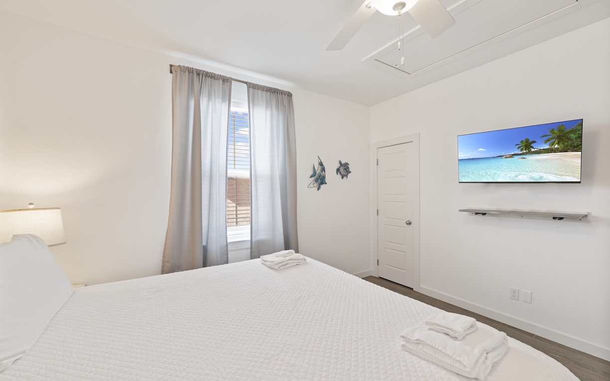 Drift off in your serene bedroom with crisp white linens, tropical wall art, and a mounted TV for relaxation.