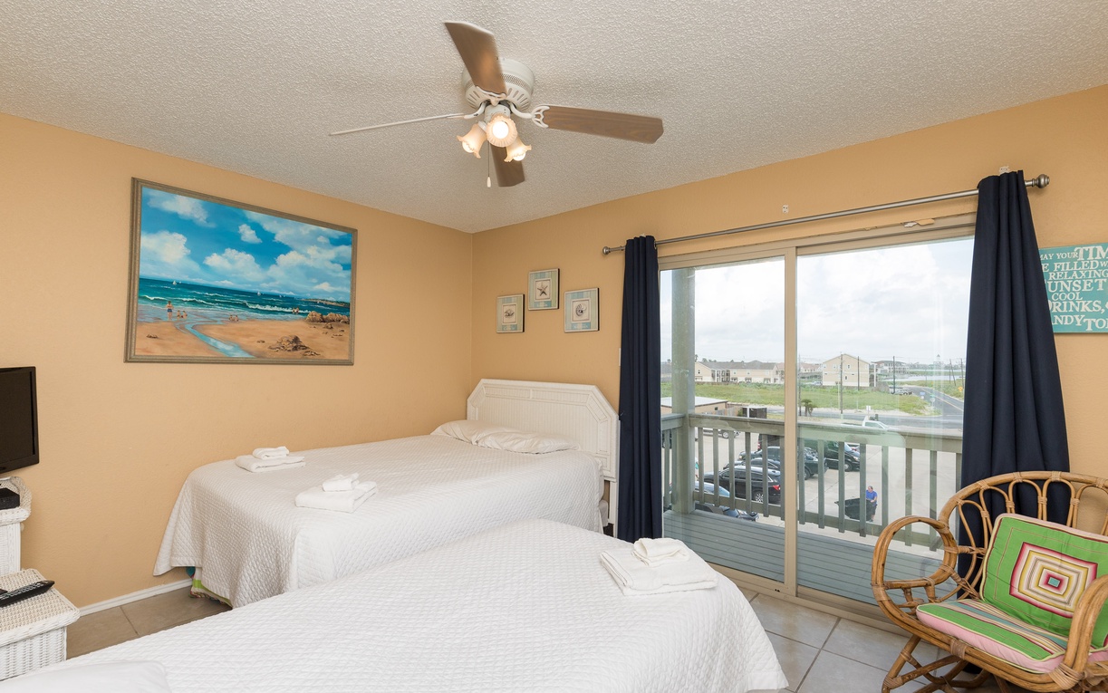 Wake up in your cozy twin bedroom with cheerful beach artwork, ceiling fan comfort, and balcony doors that flood the space with natural light.
