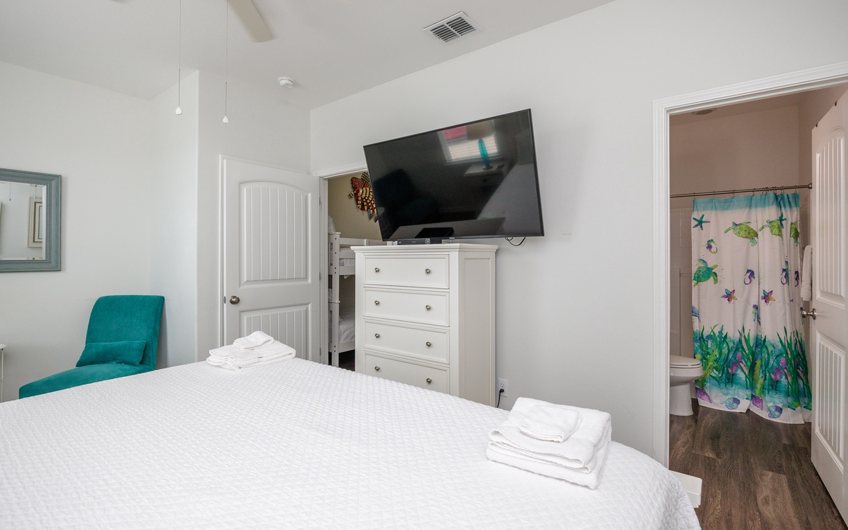 Your bright bedroom features crisp linens, a cozy teal reading chair, and wall-mounted TV for relaxing evenings after exploring.