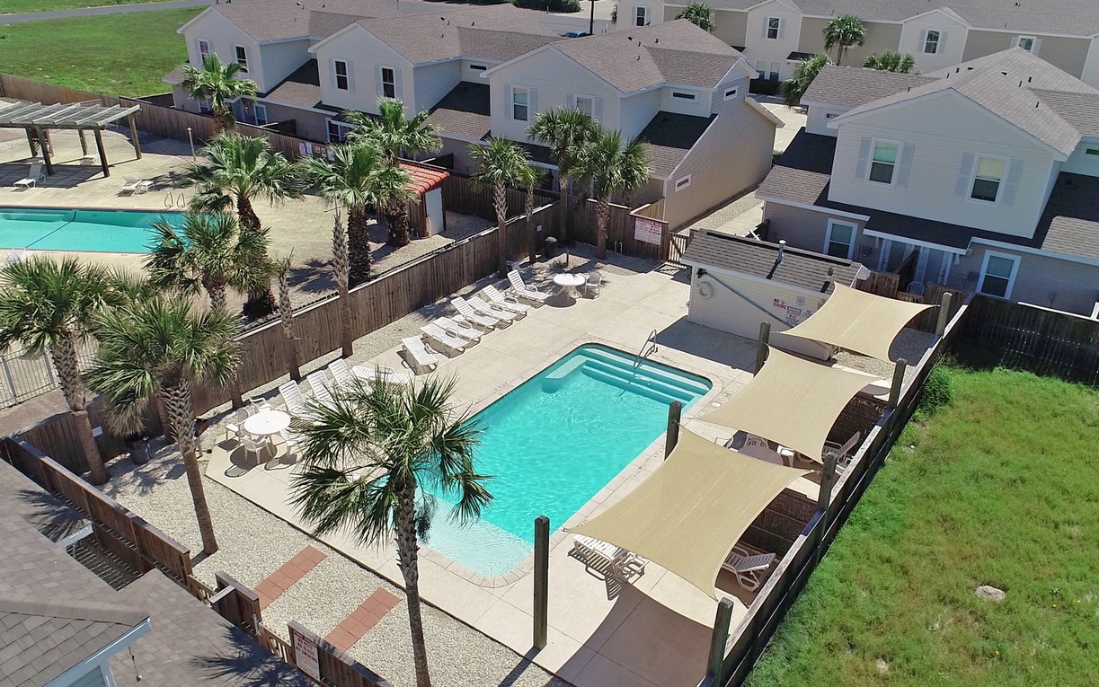 Aerial view of the property's pool area surrounded by palm trees and vacation rental homes in a peaceful residential setting.