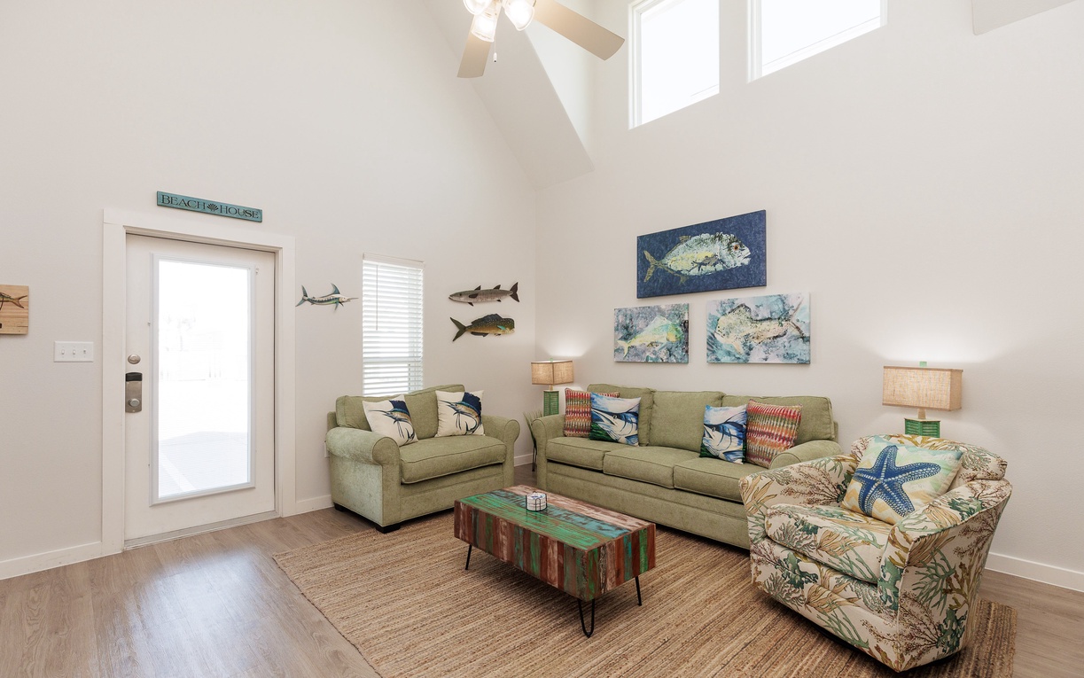Relax in your coastal-themed living room with comfortable seating, charming fish art, and soaring ceilings that create an airy, beachy atmosphere.
