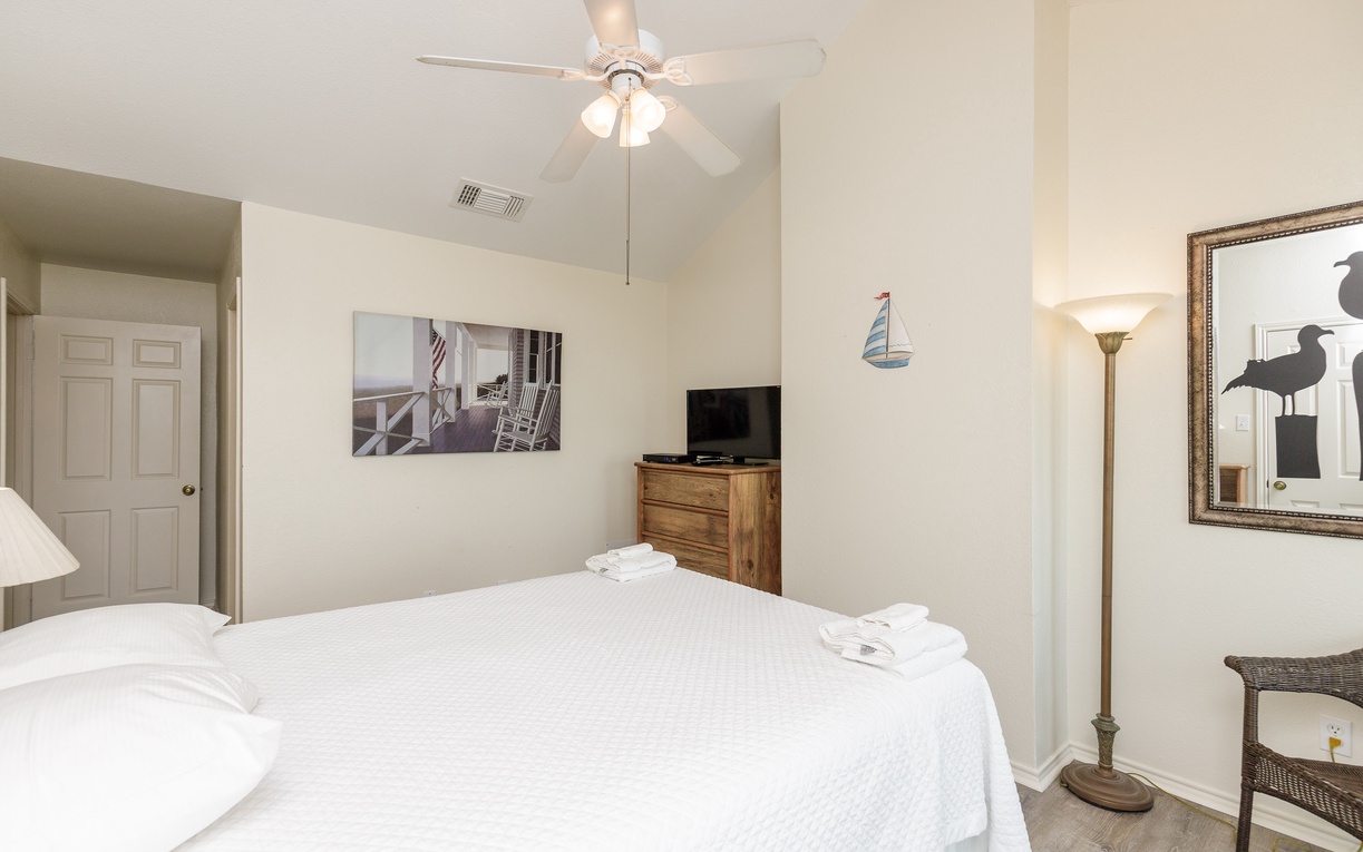 Drift off to sleep in your peaceful queen size bed in the primary bedroom, featuring fresh linens, coastal artwork, and a gentle ceiling fan breeze.