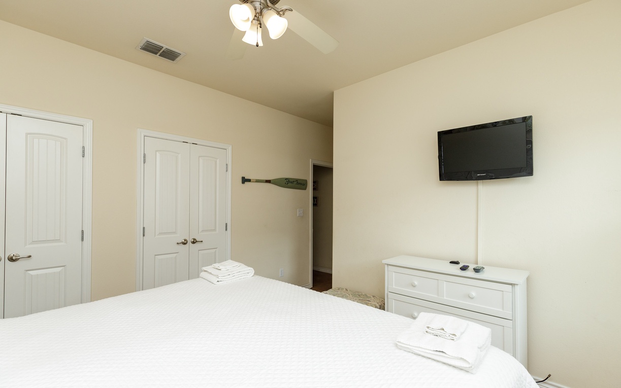 Your serene bedroom features crisp white linens, a wall-mounted TV, and bright natural light from the ceiling fan overhead.