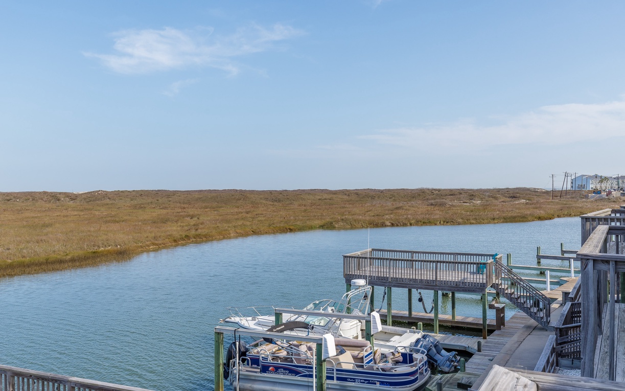 Waterfront marina with boats docked along wooden pier, surrounded by coastal marshlands under bright blue skies.