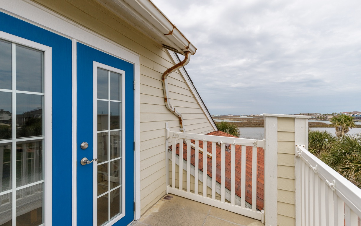 Charming coastal balcony with blue French doors opens to sweeping water views and palm trees.