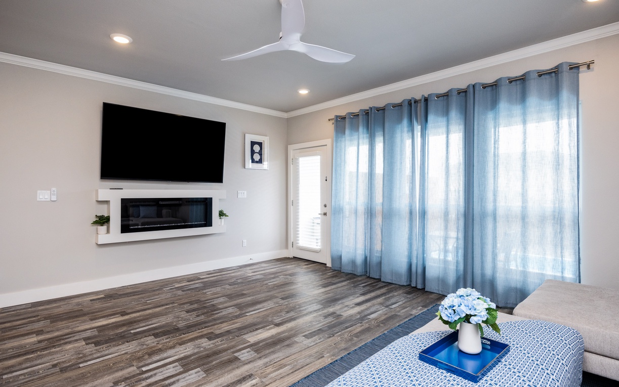 Unwind in your spacious living room where a cozy fireplace meets modern entertainment, while soft blue curtains filter gentle natural light.