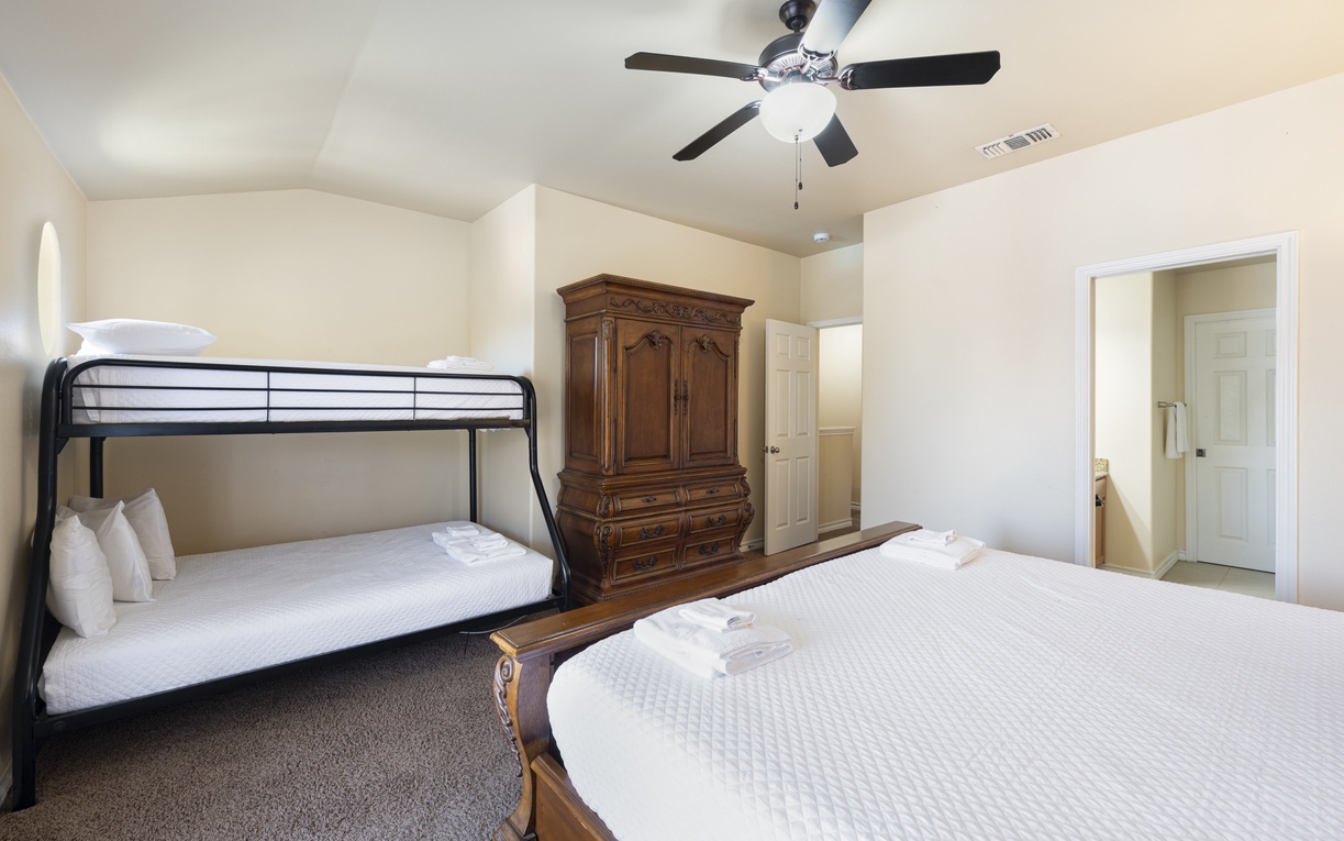 Retreat to the comfortable primary bedroom featuring a king bed, twin-over-twin bunk beds, plush bedding, a ceiling fan, a flat-screen TV, and private balcony access for a breath of fresh coastal air.