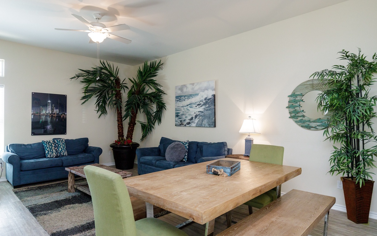 Relax in your open-concept living space with comfortable navy sofas, tropical palms, and a rustic dining table perfect for family meals.