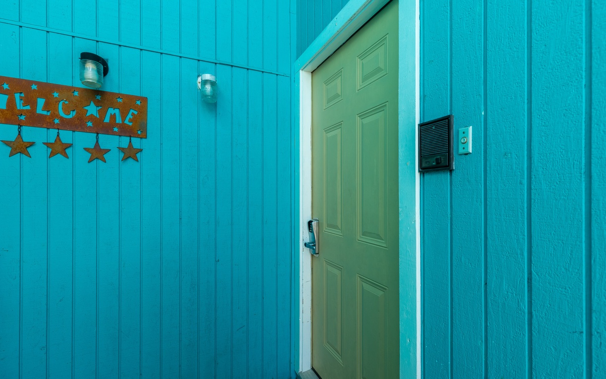Bright turquoise siding frames your welcoming entrance, where a cheerful sage door and rustic welcome sign create an instantly inviting first impression.