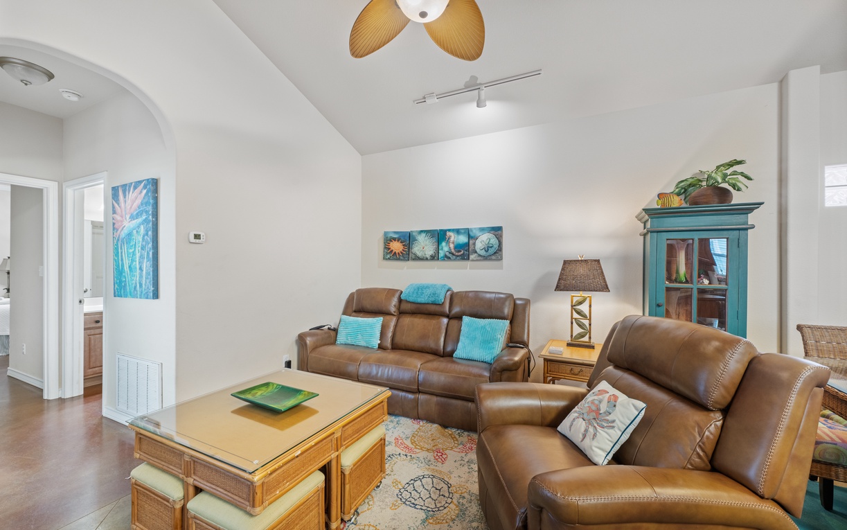 Unwind in your stylish living room with plush leather sofas, tropical ceiling fan, and coastal artwork creating a perfect beach retreat.