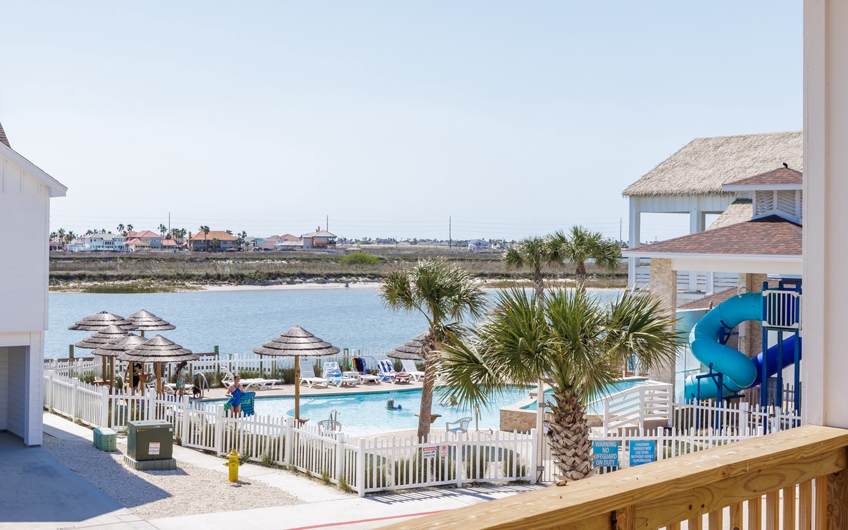Waterfront resort complex features pool area with slide, palm trees, and lagoon views in coastal community setting.