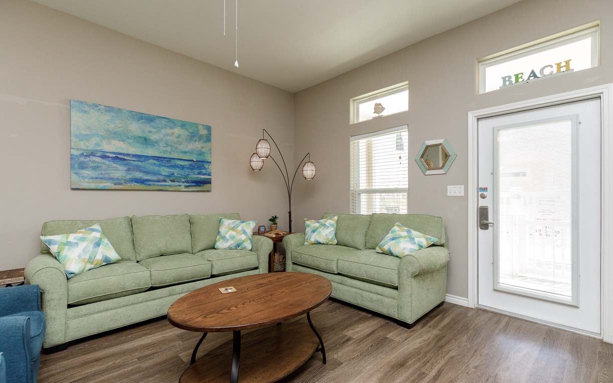 Sink into soft sage green sofas while ocean artwork sets the coastal mood in your bright, welcoming living space.