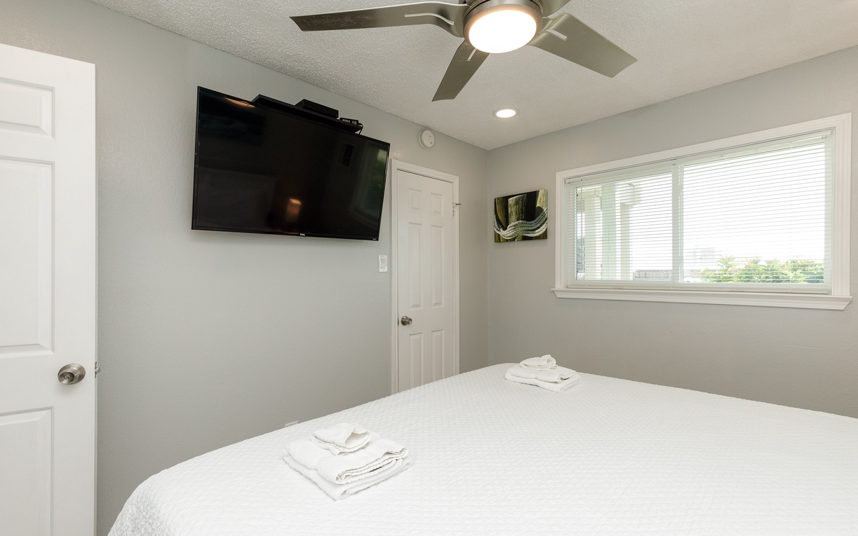 Your peaceful bedroom features crisp linens, a wall-mounted TV for relaxing evenings, and natural light streaming through blinds.