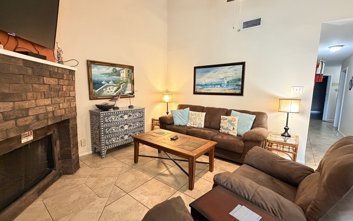 Sink into plush brown sofas by your cozy brick fireplace, where coastal artwork and warm lighting create the perfect spot for evening relaxation.