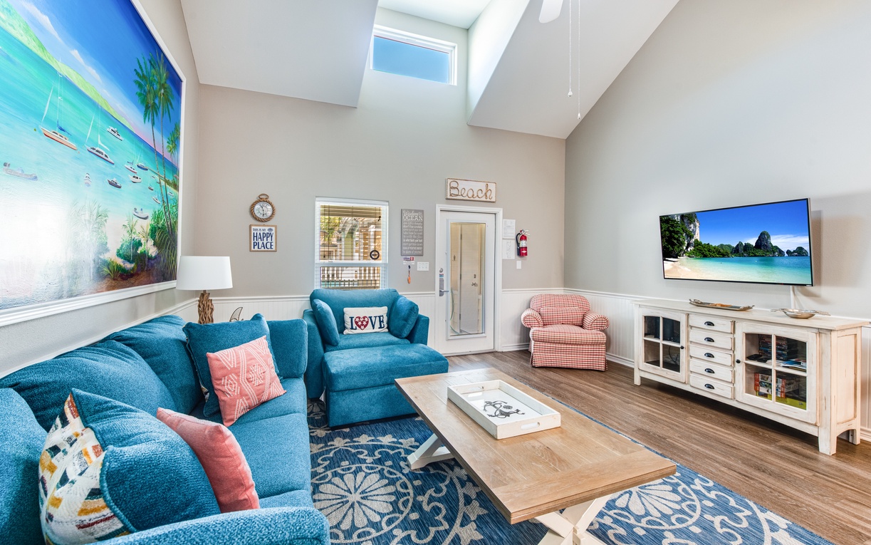 Sink into plush turquoise sofas beneath soaring ceilings and skylights, where coastal artwork and warm wood tones create your perfect beach house retreat.