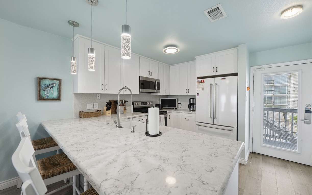 Your fully-equipped kitchen features bright white cabinetry, sleek stainless appliances, and modern pendant lighting—perfect for preparing meals while enjoying breezy coastal views from the open living area.