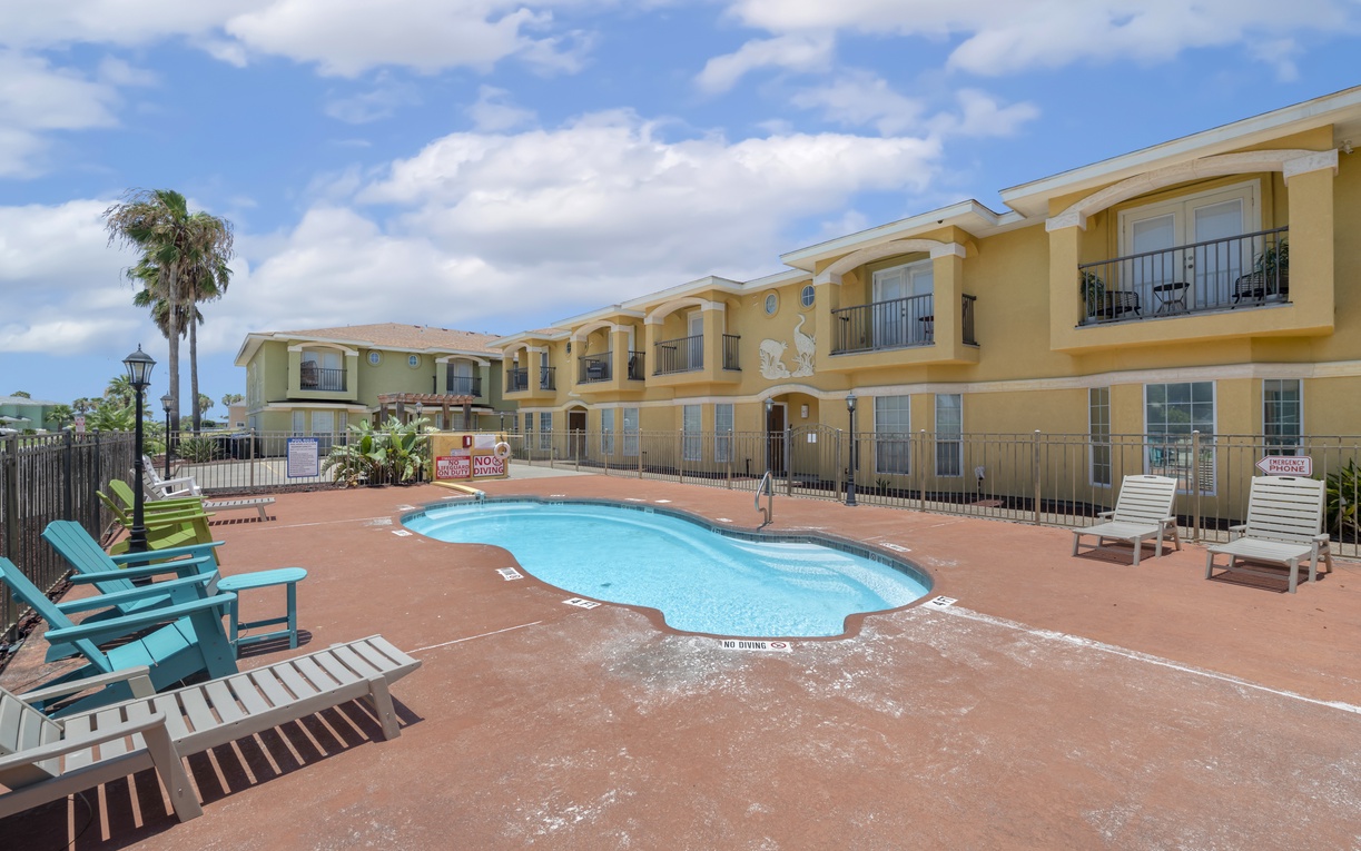 The property's welcoming courtyard features a refreshing pool surrounded by sun loungers and colorful seating, framed by tropical palms and balconied accommodations under bright coastal skies.