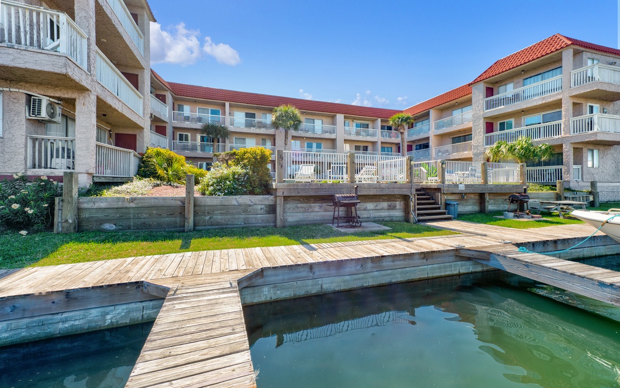 Resort-style property featuring multi-story buildings with private balconies surrounding tranquil waterfront areas and wooden walkways under bright blue skies.
