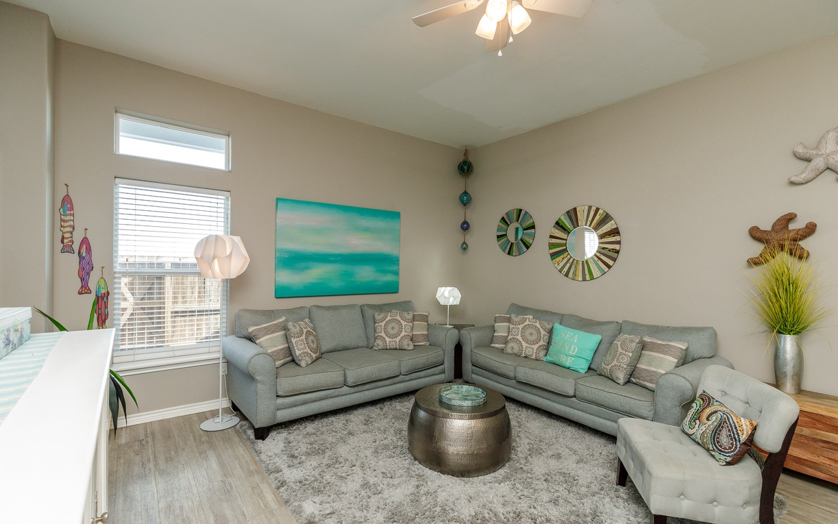 Unwind in your stylish living room featuring plush gray sectional seating, coastal artwork, and warm lighting that creates the perfect atmosphere for relaxation.