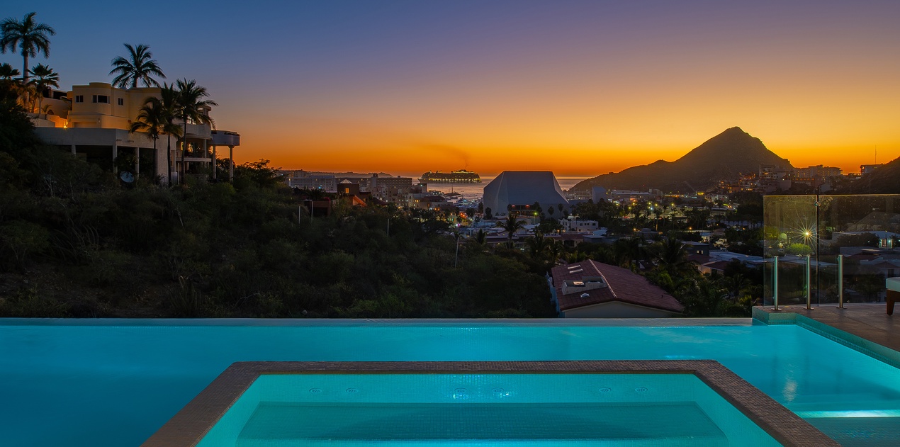 Stunning infinity pool overlooking tropical coastline at sunset, with dramatic mountain silhouettes and vibrant orange-purple sky creating the perfect evening backdrop.