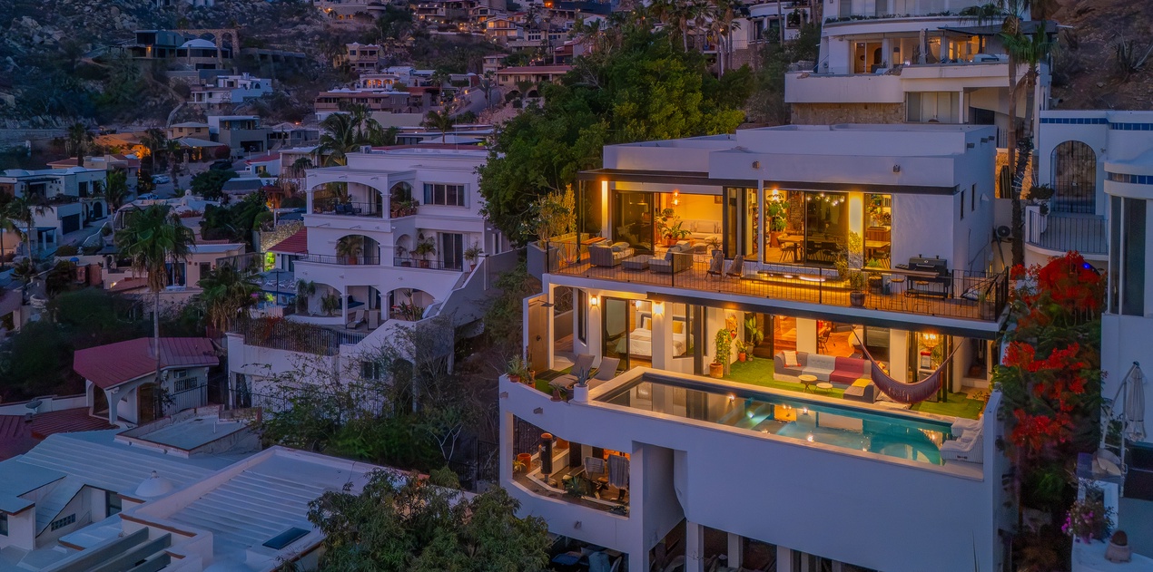 Stunning hillside property nestled in a vibrant coastal neighborhood, featuring multiple terraces and a pool with warm evening lighting illuminating the modern architecture.
