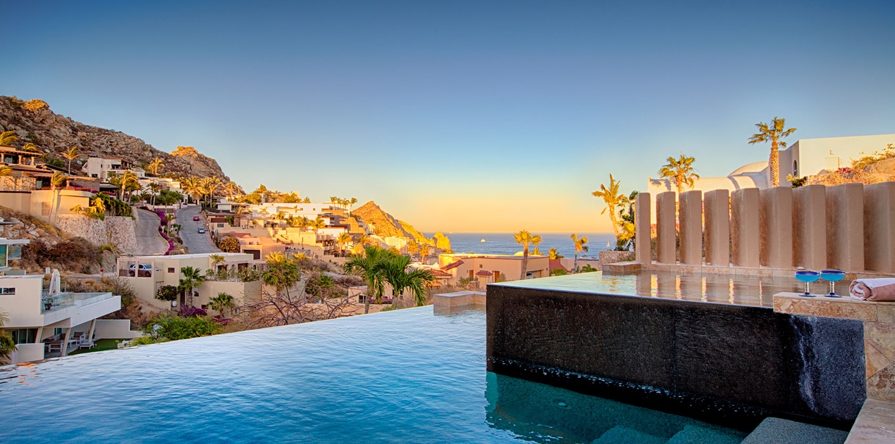 An infinity pool overlooking a hillside coastal community during golden hour. The property sits among terraced homes carved into the rocky landscape, with ocean views stretching to the horizon.