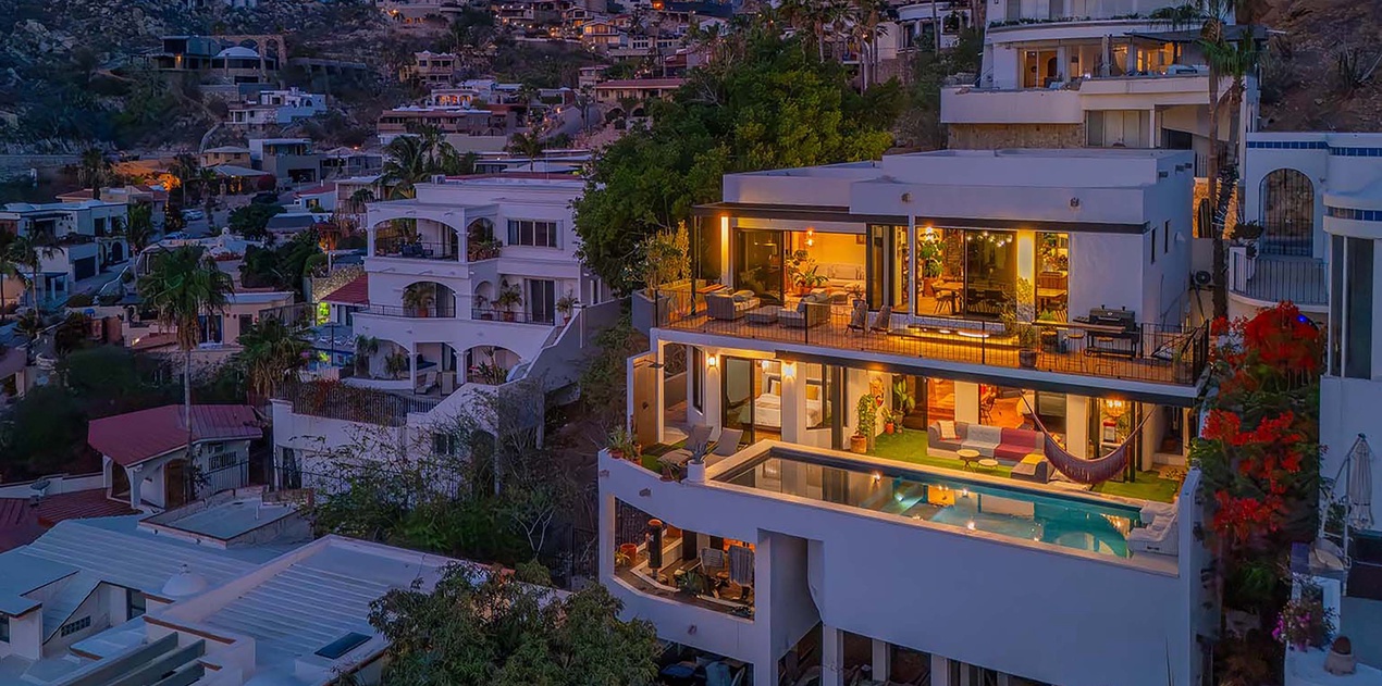 Hillside luxury villa with illuminated terraces and infinity pool at twilight, overlooking the neighboring coastal community and surrounding landscape.