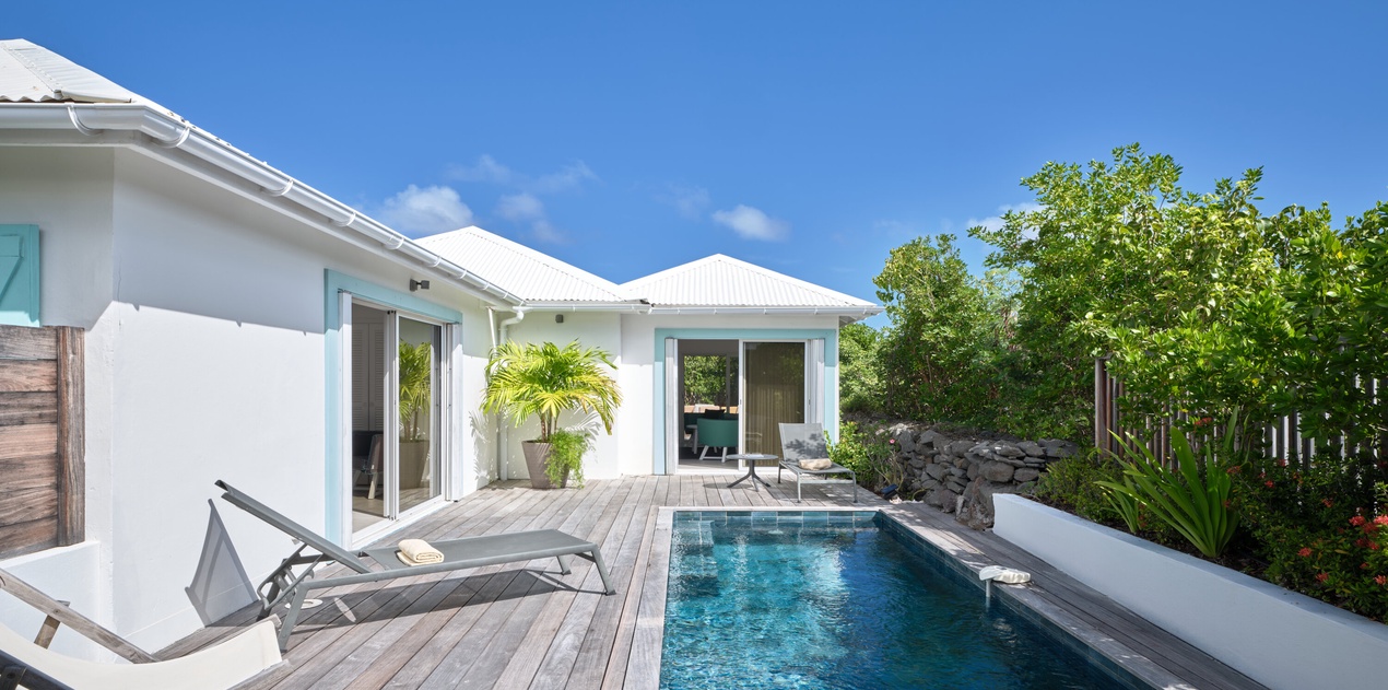 Modern white villa with private swimming pool surrounded by wooden decking and lush tropical landscaping under clear blue skies.