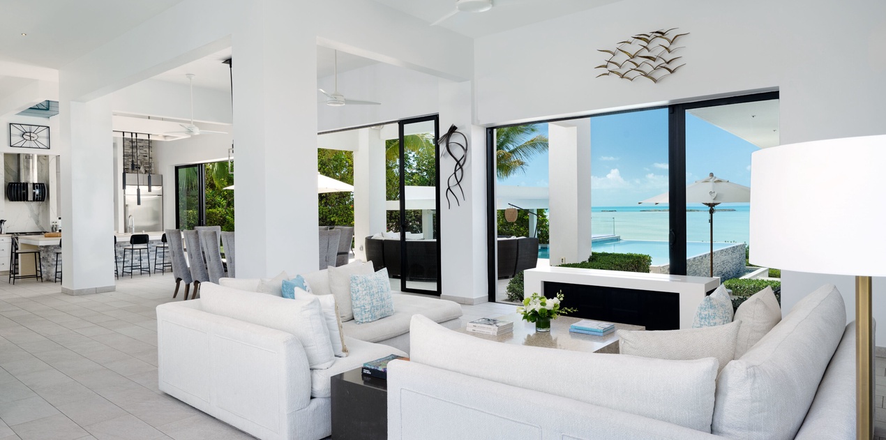 Sink into plush white sofas while turquoise waters and swaying palms frame your perfect tropical escape through floor-to-ceiling windows.
