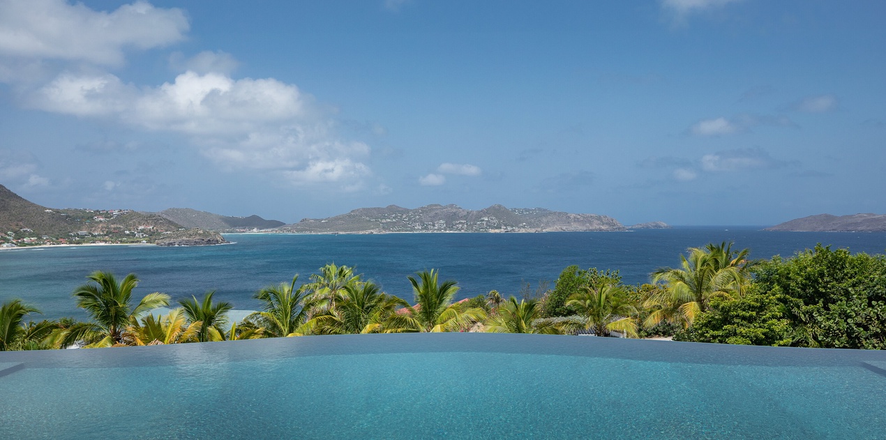 An infinity pool frames sweeping views of turquoise waters, lush palms, and distant coastal mountains under open Caribbean skies.