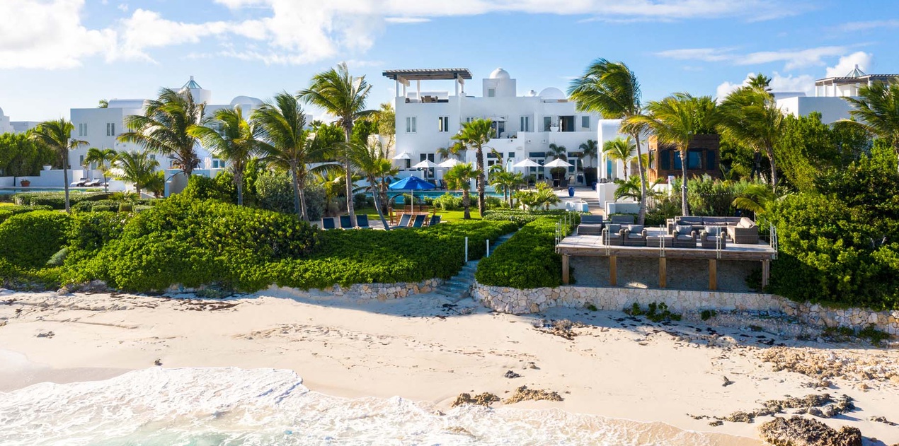 Stunning beachfront estate with white architecture, lush tropical gardens, and turquoise waters meeting sandy shores under bright blue skies.