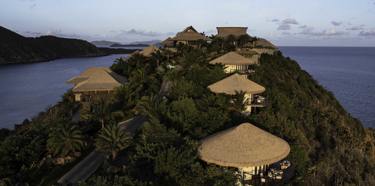 Tropical resort buildings perched dramatically on a lush hillside peninsula, surrounded by pristine blue waters and natural vegetation.