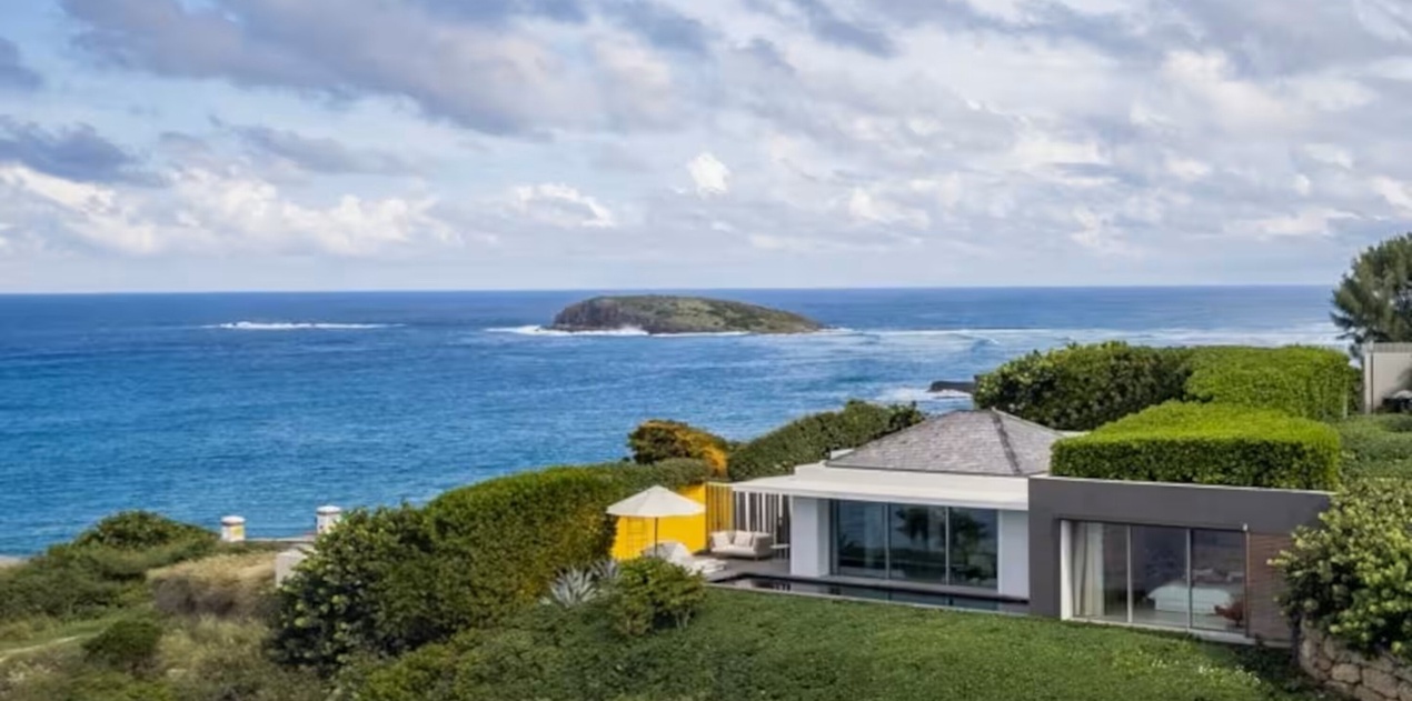 This stunning modern property sits on a coastal hillside overlooking the sparkling ocean, with a private island visible just offshore and lush native landscaping framing the sleek architecture.