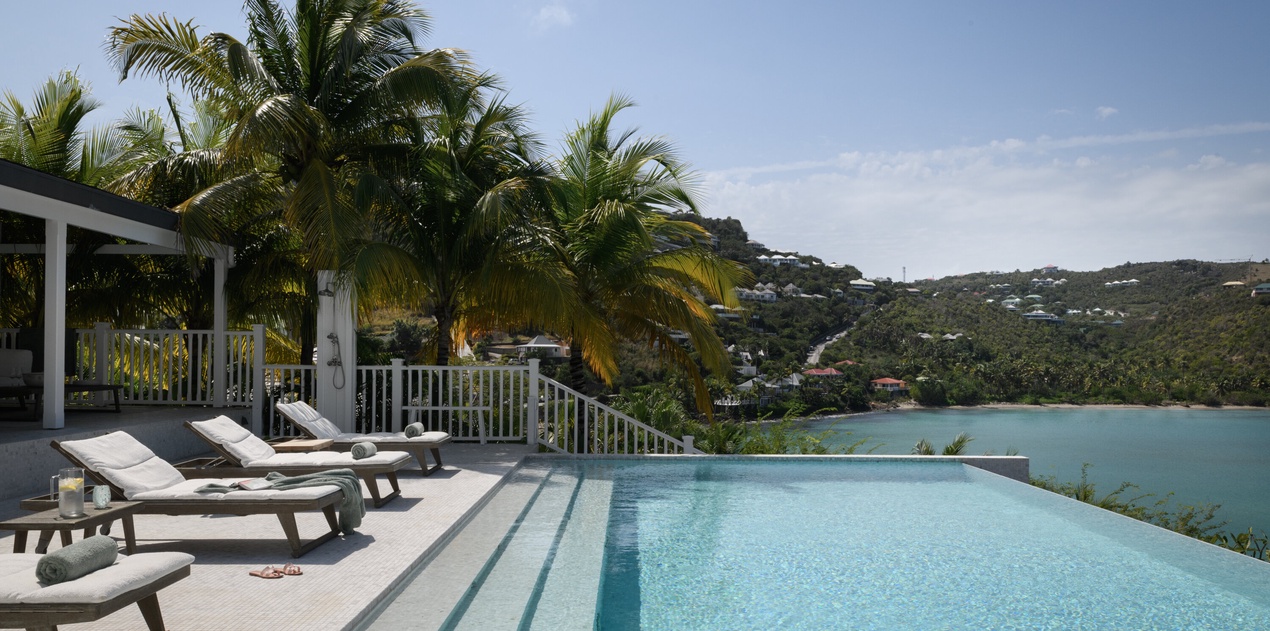 Stunning infinity pool overlooking turquoise bay waters, framed by swaying palms and hillside homes in this tropical paradise setting.