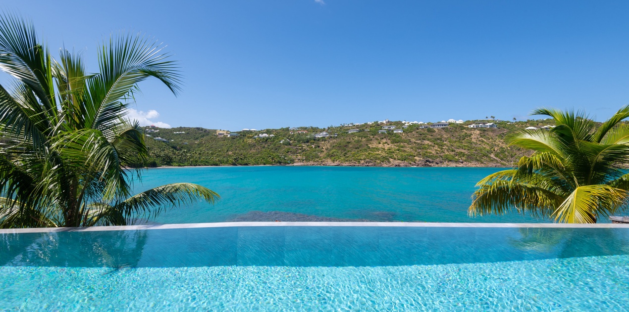 A stunning infinity pool creates the illusion of merging with crystal-clear turquoise waters beyond, framed by swaying palm trees.