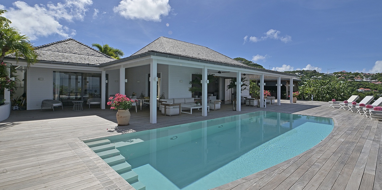 A contemporary villa showcases its sleek covered terrace and pristine swimming pool, surrounded by tropical landscaping under bright skies.