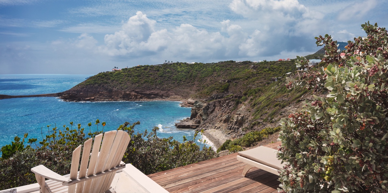 A secluded coastal hillside offers a dramatic panoramic view of turquoise waters, rugged cliffs, and lush green vegetation stretching toward distant villas under a Caribbean sky.