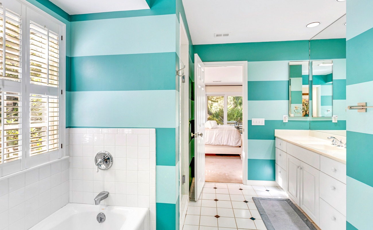 Refresh in your vibrant turquoise-striped bathroom with white tiled tub, dual vanity, and tropical garden views through plantation shutters.