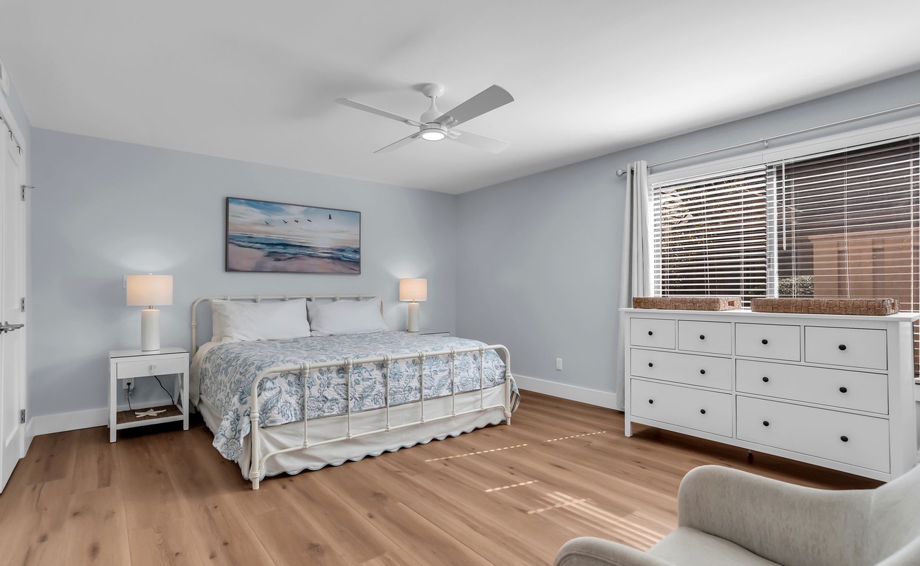 A serene primary bedroom retreat featuring a plush king bed dressed in soft coastal linens and soothing neutral tones. Light wood flooring and clean white furnishings create a bright, airy atmosphere perfect for restful nights.