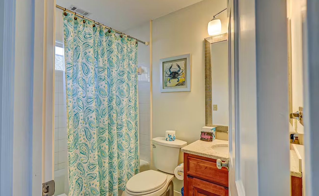 Refresh in your private bathroom with coastal charm—paisley curtain, warm wood vanity, and nautical artwork create a welcoming retreat.