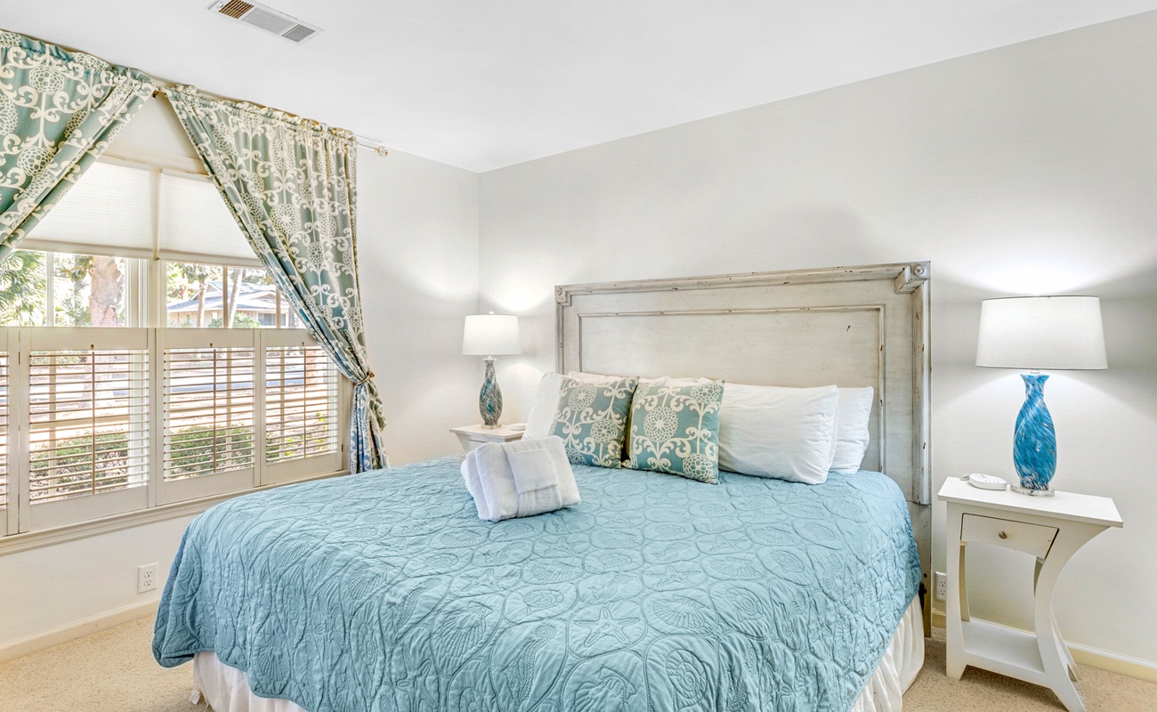 Drift off in this serene bedroom with soft blue linens, natural light streaming through plantation shutters, and coastal-inspired décor.