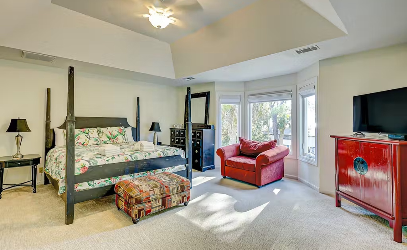 Unwind in your spacious master suite featuring elegant four-poster bed, cozy reading chair, and tropical garden views through expansive windows.