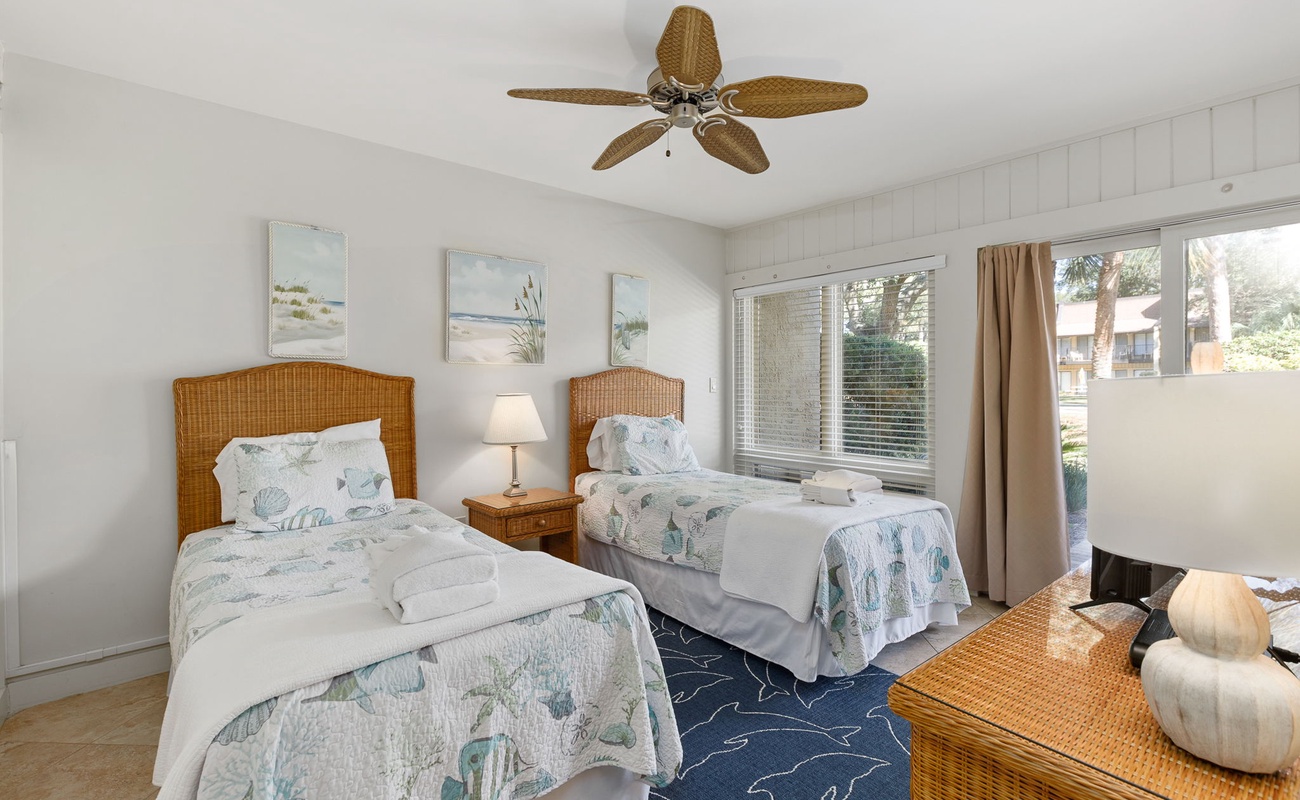 Relax in this coastal twin bedroom with wicker furniture, ocean-inspired bedding, and a cooling ceiling fan for your comfort.