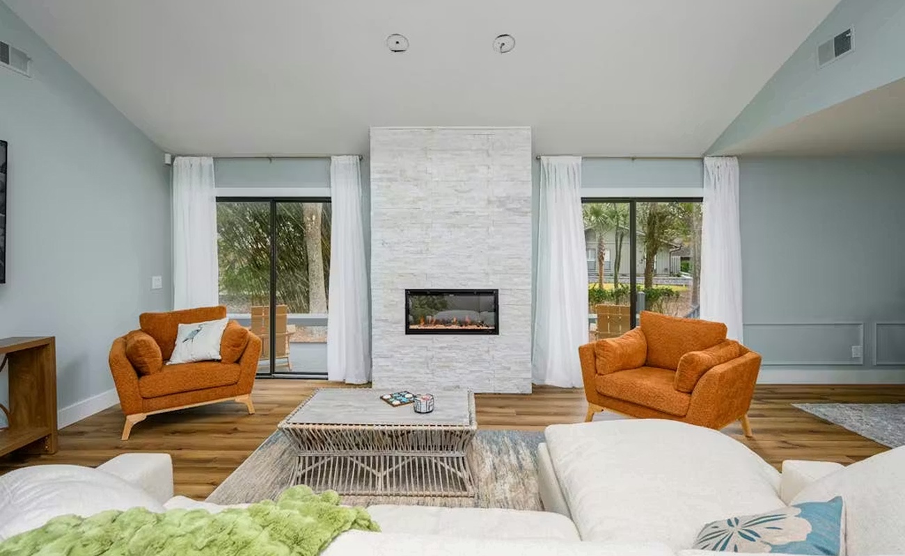 Relax in your stylish living room with modern fireplace, cozy orange armchairs, and peaceful garden views through floor-to-ceiling windows.