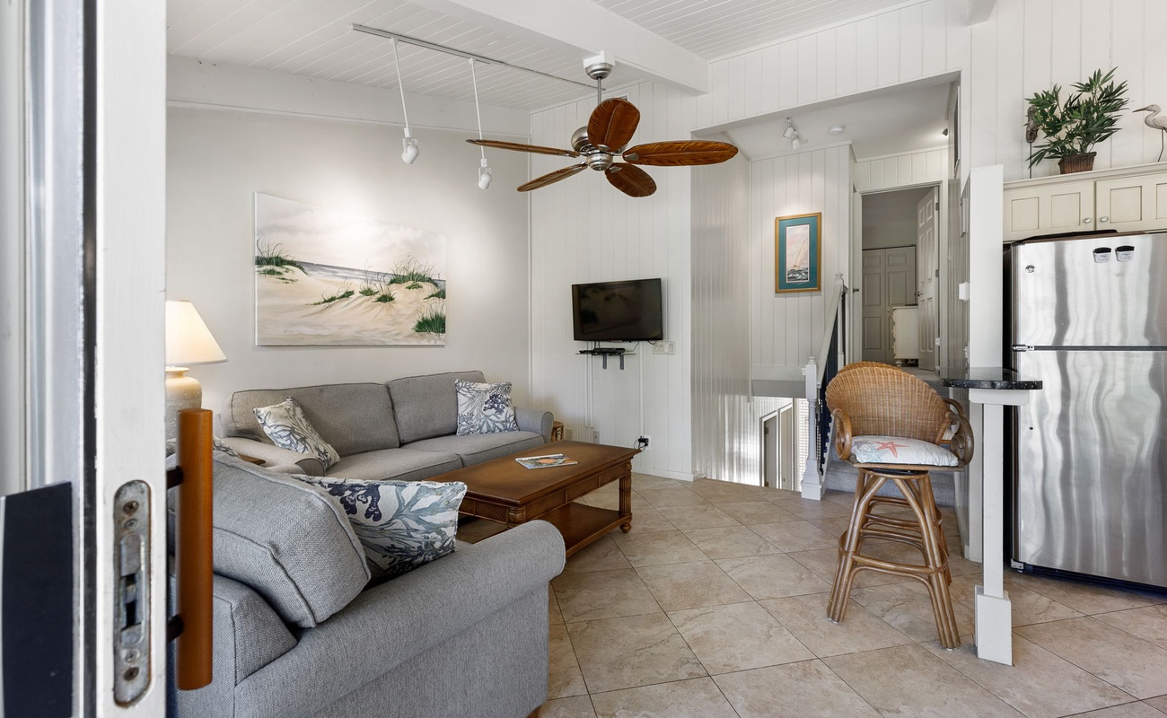 Sink into the cozy sectional and enjoy your coastal retreat with ocean-themed art, tropical ceiling fan, and modern kitchen nearby.
