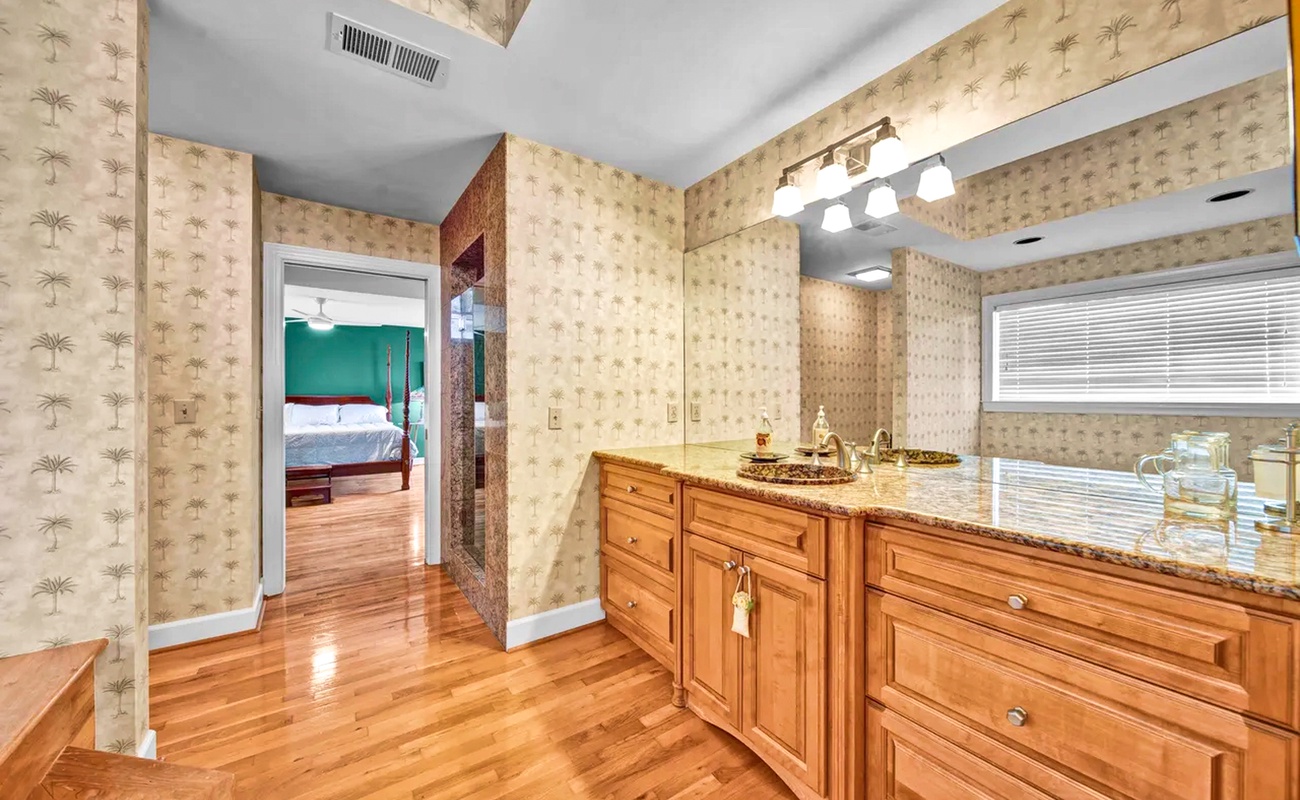 Your elegant en-suite bathroom features warm wood vanity, granite counters, and charming palm-patterned wallpaper creating a tropical oasis.