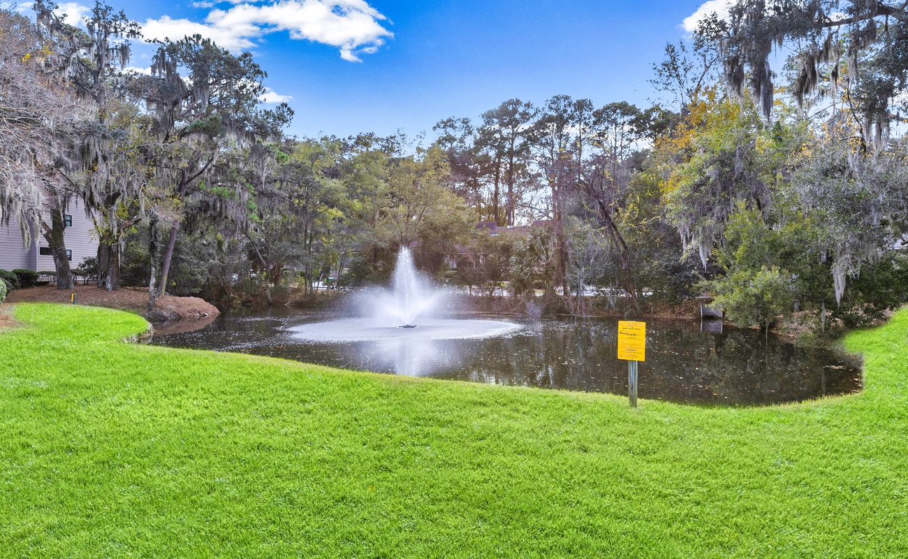 Peaceful pond with fountain surrounded by lush green space and mature trees draped with Spanish moss.