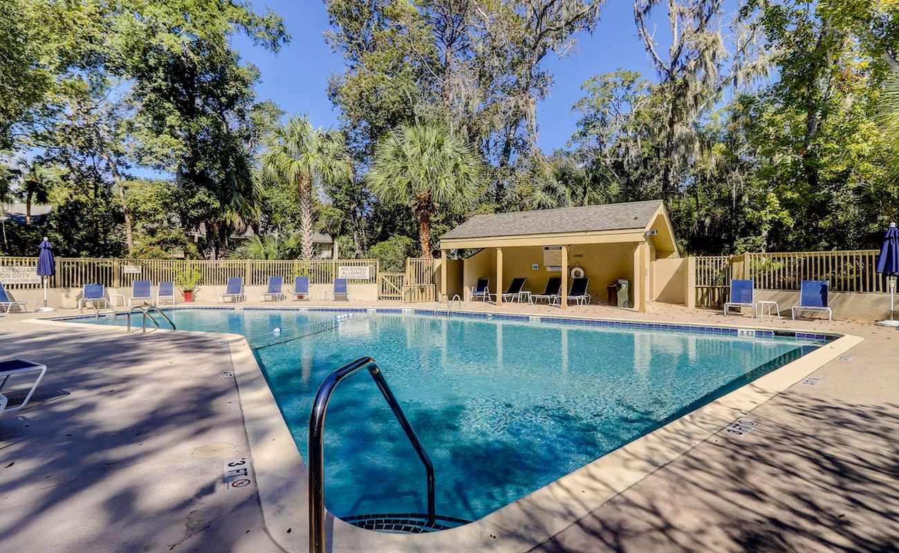 A sparkling outdoor pool area surrounded by mature trees and tropical palms, featuring a covered pavilion with seating.