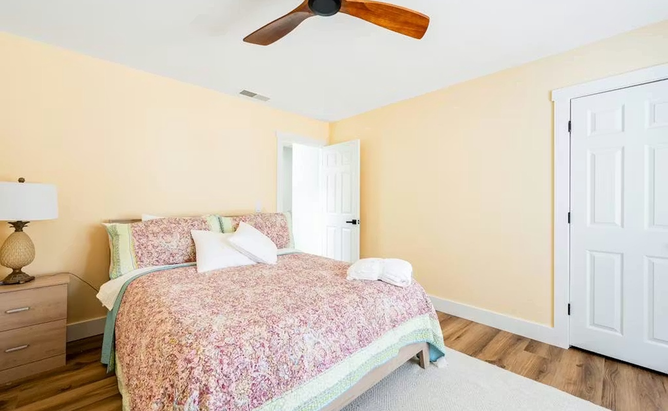 Your cozy bedroom features a comfortable bed with floral linens, warm yellow walls, and a ceiling fan for comfort.