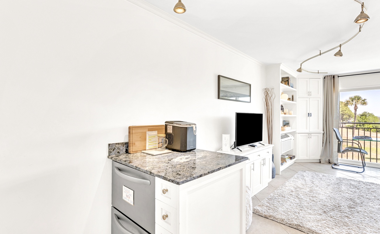 Your bright kitchenette with granite counters and modern appliances opens to a cozy living space with balcony views of palm trees.